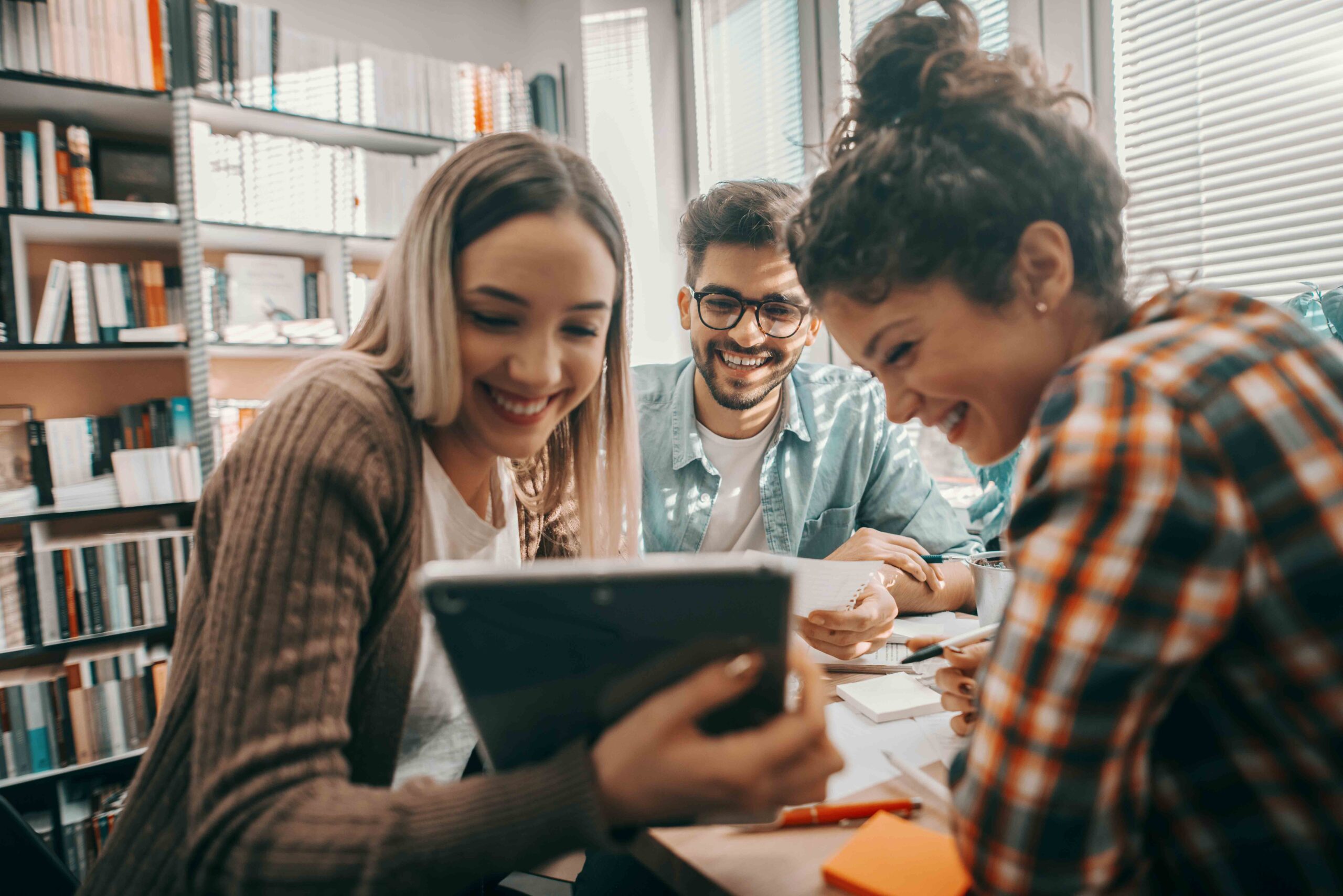 Drei glückliche Schüler in Freizeitkleidung benutzen ein Tablet für ein Schulprojekt und sitzen am Schreibtisch in der Bibliothek.