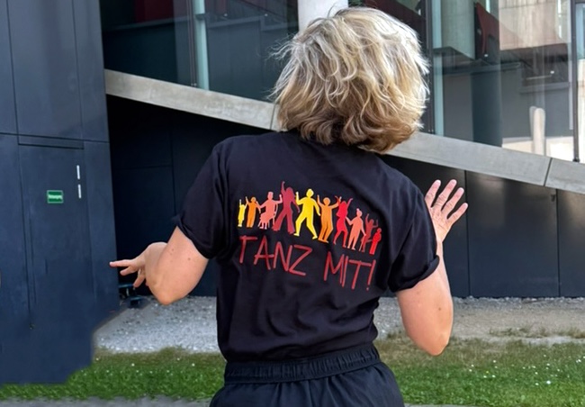 Eine Rückensicht einer vermutlich weibliche Person. Sie tanzt und trägt ein schwarzes T-Shirt mit dem Aufdruck des Projektlogos.