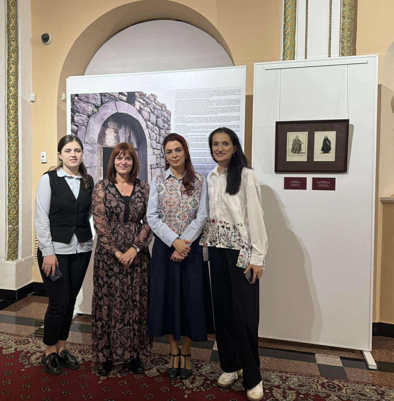 Eine Gruppe von vier Frauen vor Leinwänden, die armenische Ausstellungsstücke zeigen.