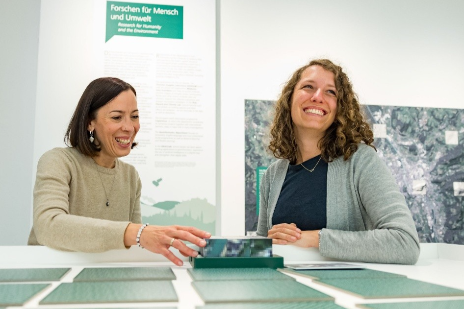 Zwei lachende Frauen sitzen vor einem Rollup an einem Tisch, auf dem grüne Kärtchen liegen. Im Hintergrund ist ein Satellitenbild zu sehen.