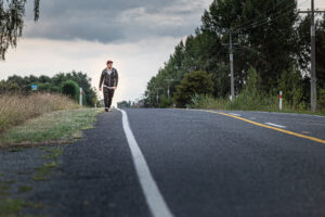Eine Person geht eine verlassene Straße entlang. Seitlich ist Wald zu sehen.