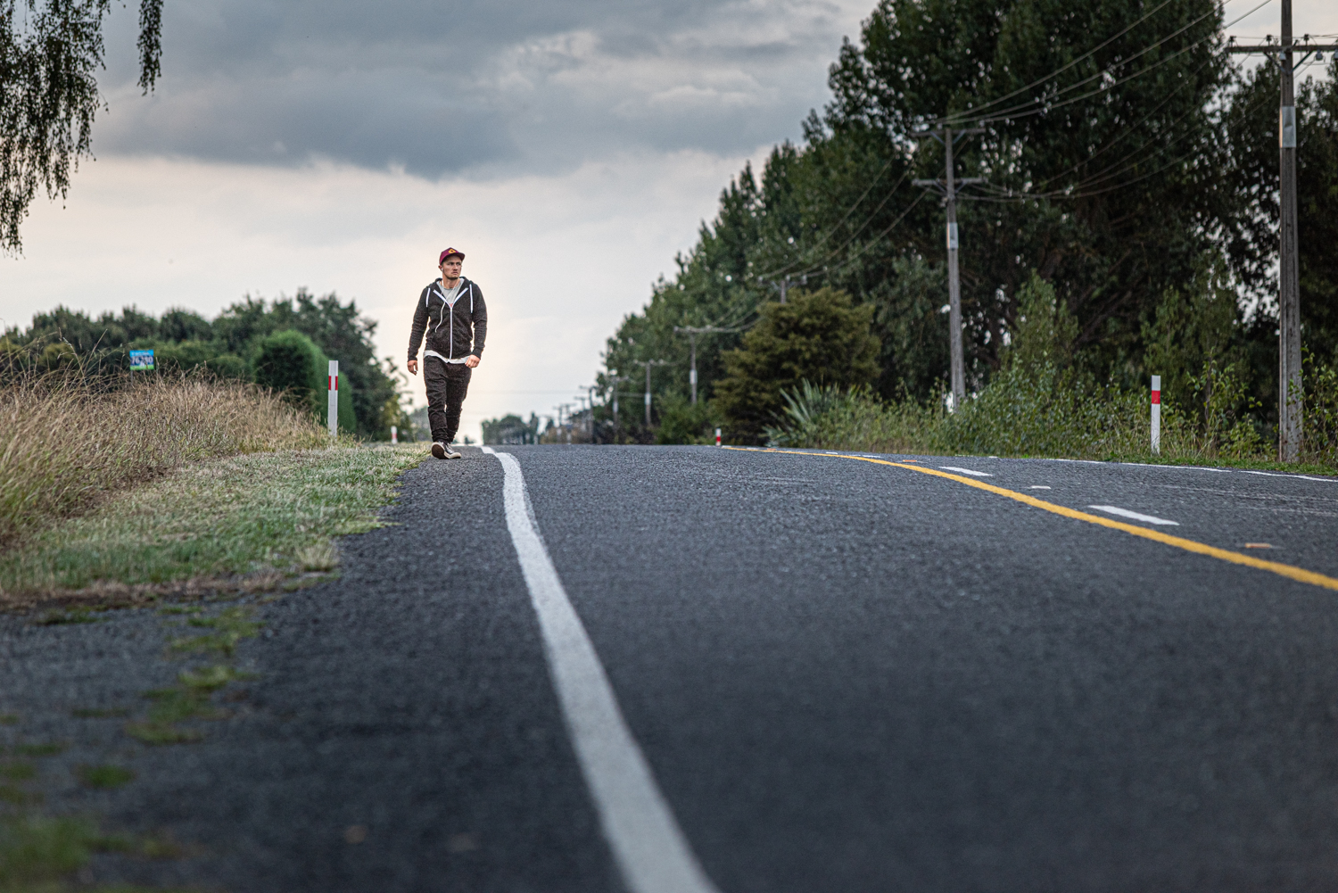 Ein querformatiges Bild, das eine einsame Straße zeigt, die durch eine waldige Gegen führt. Am Horizont ist eine Person zu sehen, die zu Fuß geht.