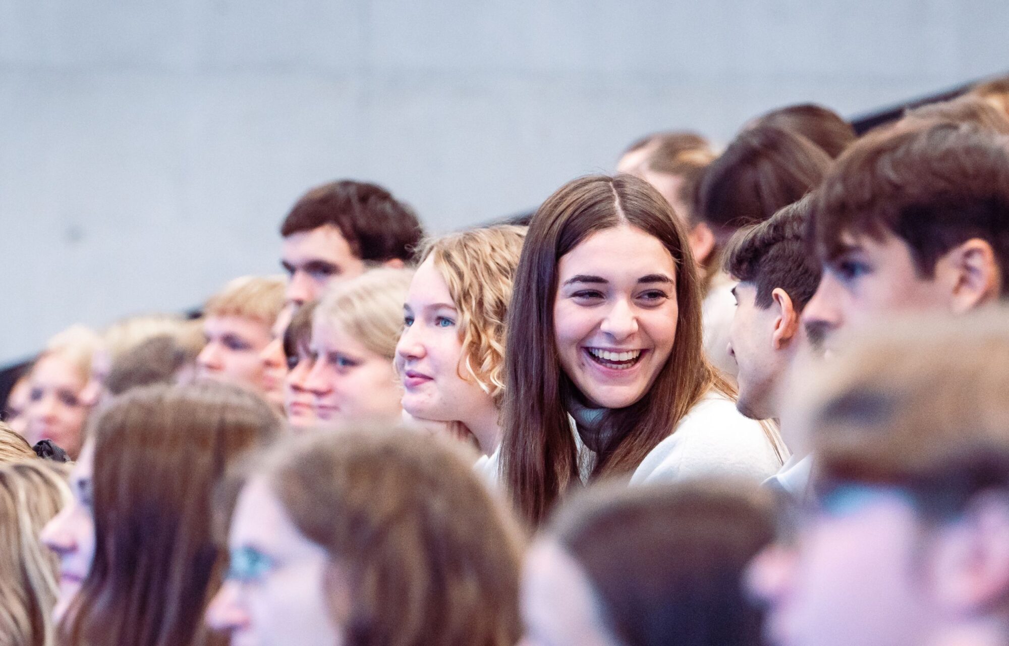 Studentin sitzt mit anderen Studierenden im H&ouml;rsaal und lacht in die Kamera