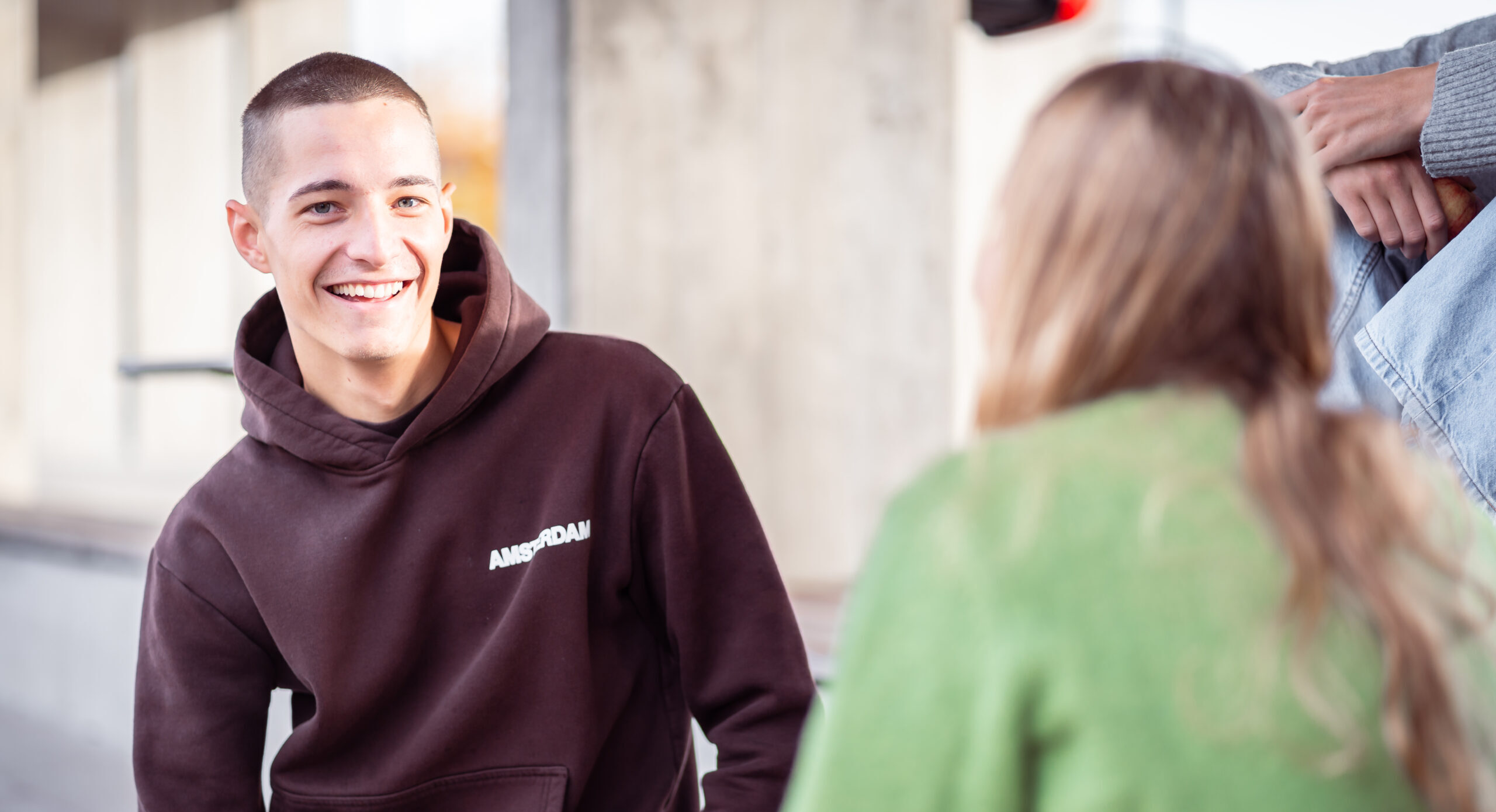 Fokus auf l&auml;chelnden jungen Mann, der sich mit einer jungen Frau unterh&auml;lt, die mit dem R&uuml;cken zur Kamera sitzt.
