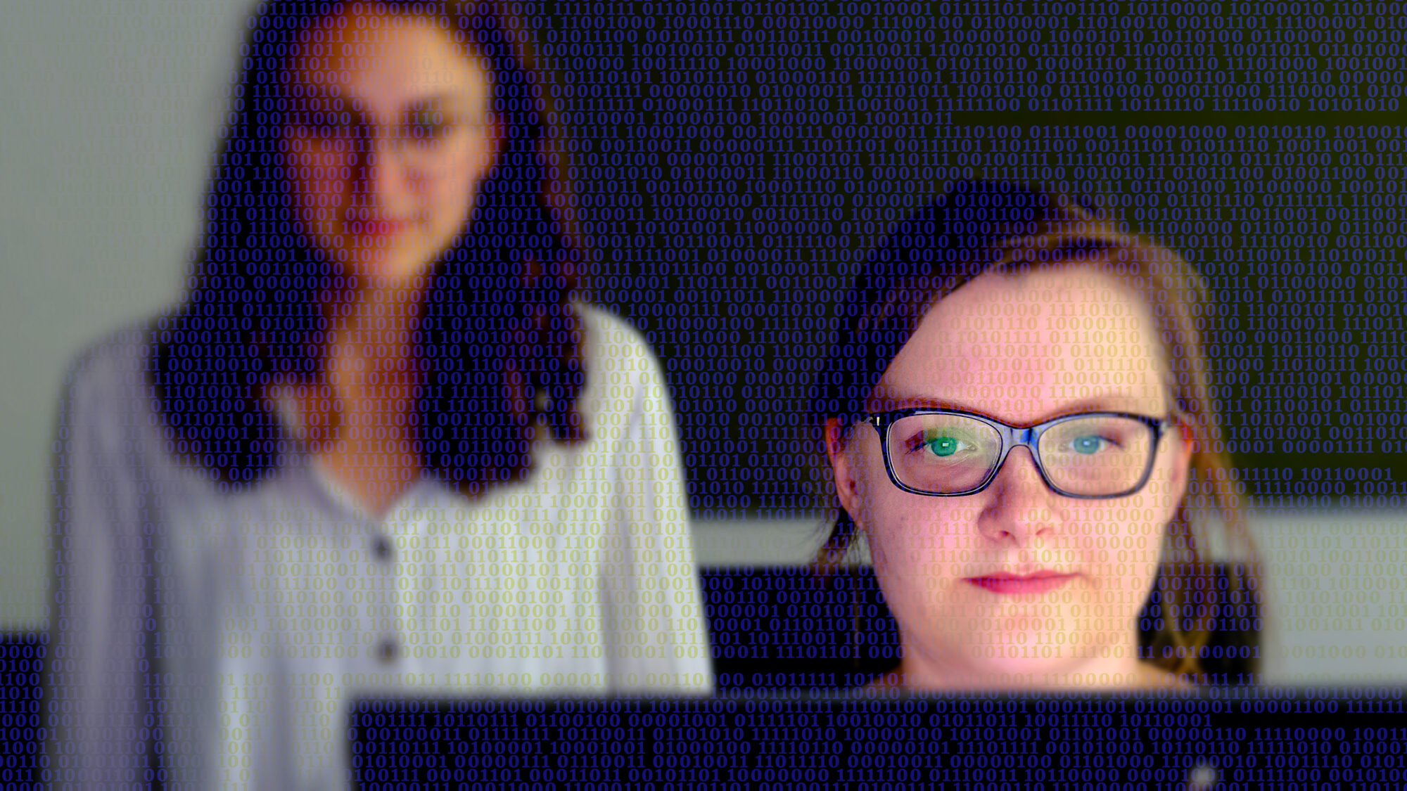 Frau sitzt vor Bildschirm, der ihr Gesicht beleuchtet. Im Hintergrund blickt eine weitere Frau auf den Bildschirm. Über dem gesamten Bild verlaufen Zahlencodes.